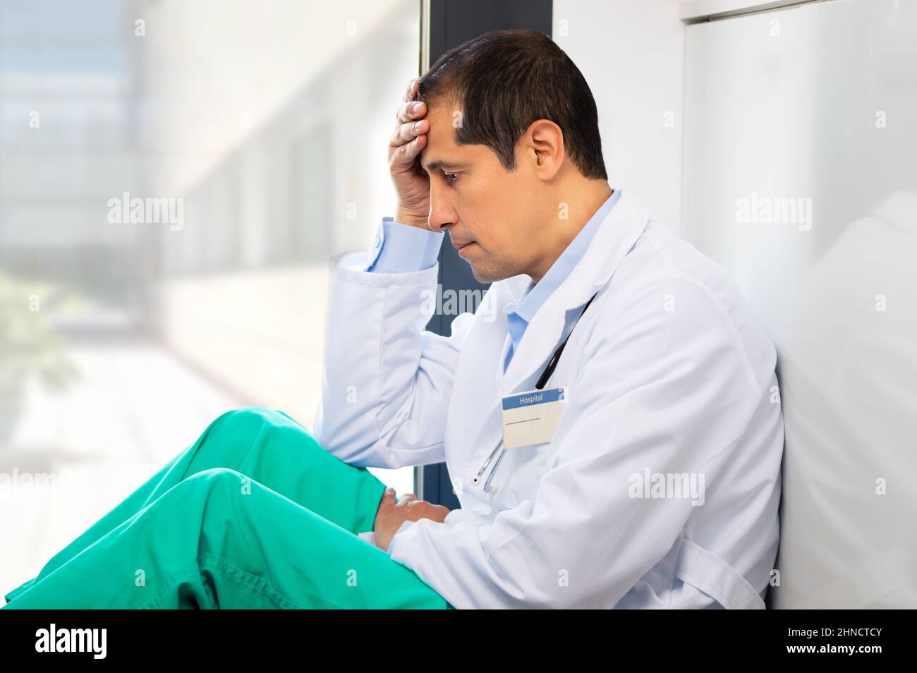 Plan court d'un jeune homme beau docteur avec un mal de tête tenant sa tête tout en s'appuyant contre un mur dans son hôpital Banque D'Images