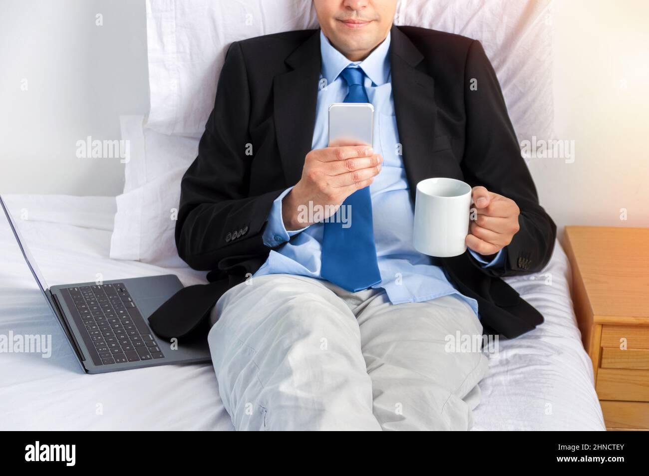 Homme d'affaires mature tenant une tasse de café et travaillant avec un smartphone sur le lit Banque D'Images