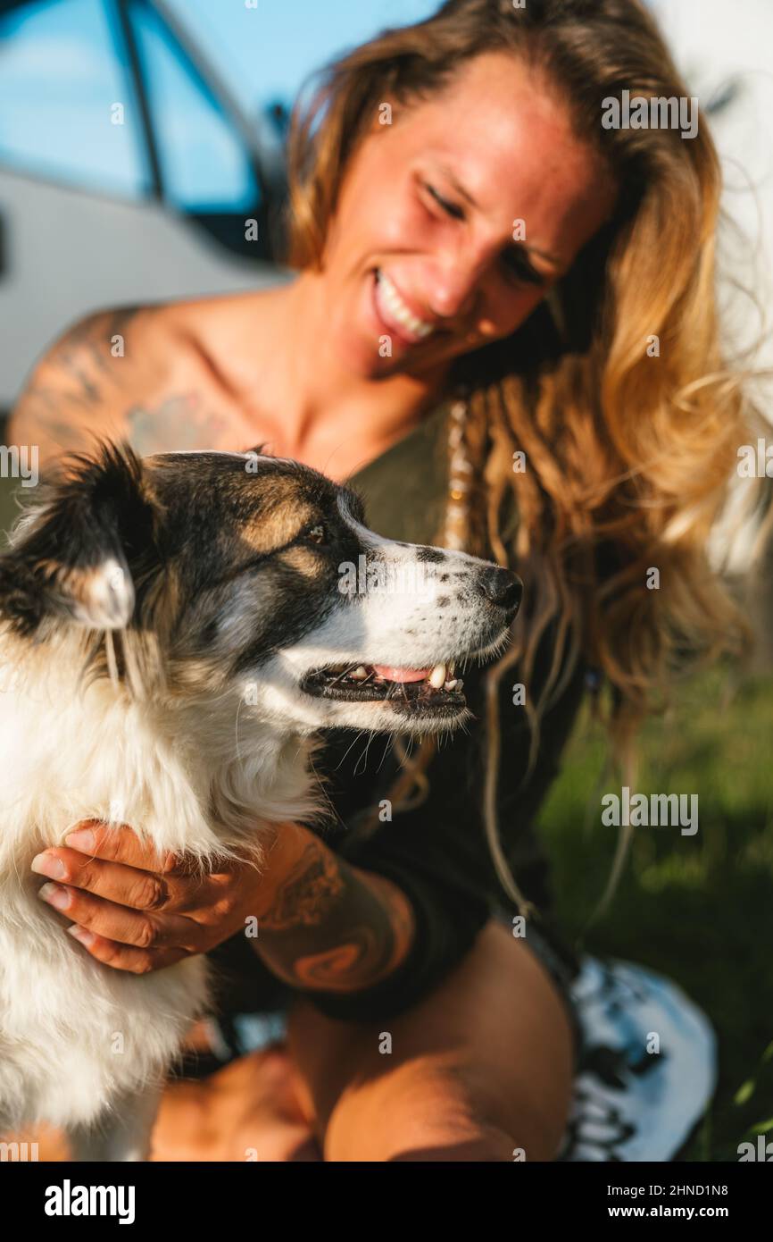Une femme positive qui embrasse un chien de berger australien coiffé avec un museau à pois tout en étant assise dans une rue ensoleillée le jour de l'été Banque D'Images
