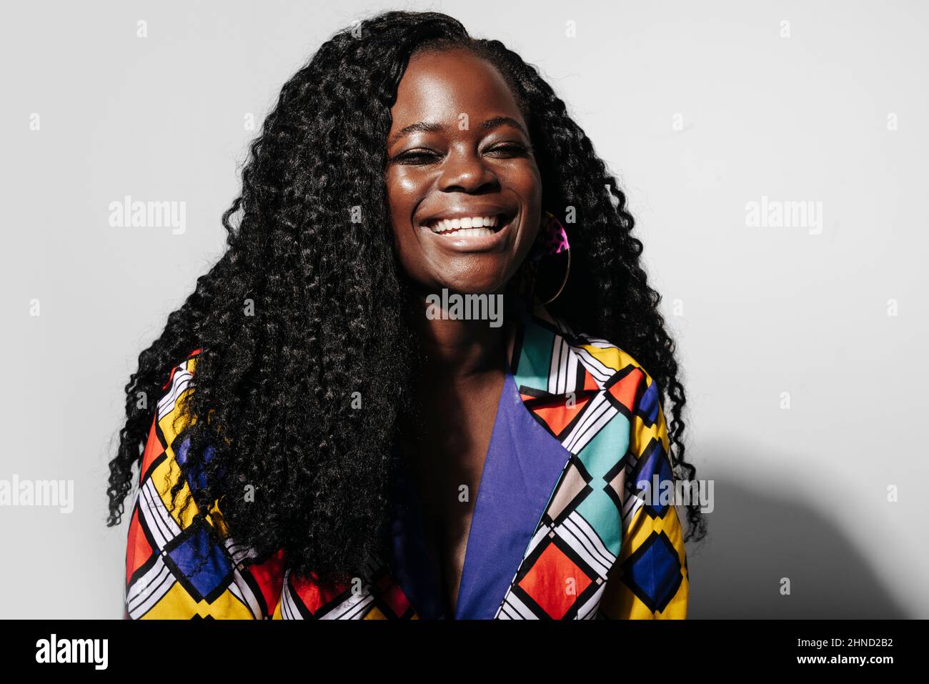 Bonne femme afro-américaine avec des cheveux bouclés souriant à la caméra avec les yeux fermés tout en se tenant contre le mur blanc Banque D'Images