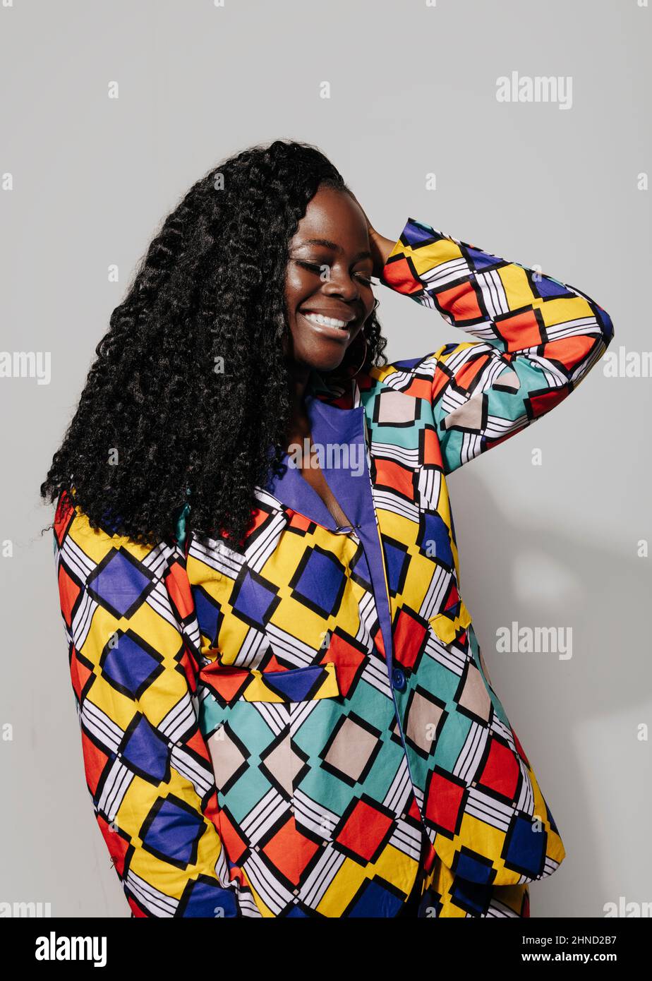 Bonne femme afro-américaine aux cheveux bouclés dans une tenue élégante en souriant sur fond blanc avec main sur la tête Banque D'Images
