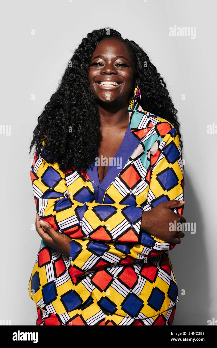 Bonne femme afro-américaine avec des cheveux bouclés dans une tenue élégante souriant et regardant l'appareil photo tout en se tenant debout sur fond blanc Banque D'Images
