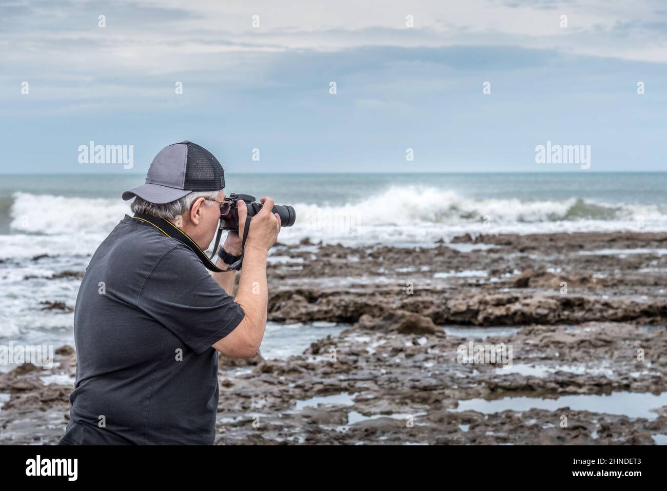 Vue arrière du demi-corps d'un homme adulte adulte adulte adulte avec Cap prenant une photo sur les rochers près de la mer. Banque D'Images