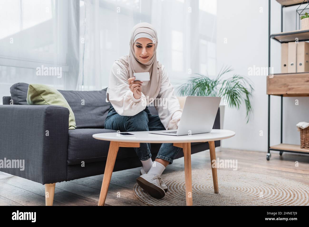 femme arabe souriante regardant la carte de crédit tout en étant assise sur un canapé près d'un ordinateur portable Banque D'Images
