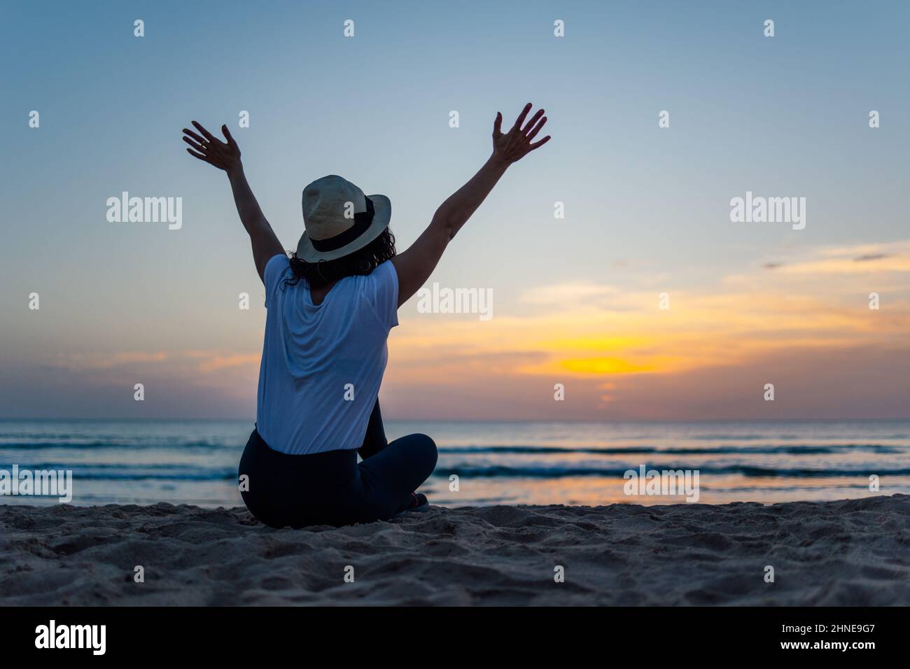 Jeune femme méconnaissable en chapeau levant les bras excitée par le coucher de soleil sur la plage Banque D'Images