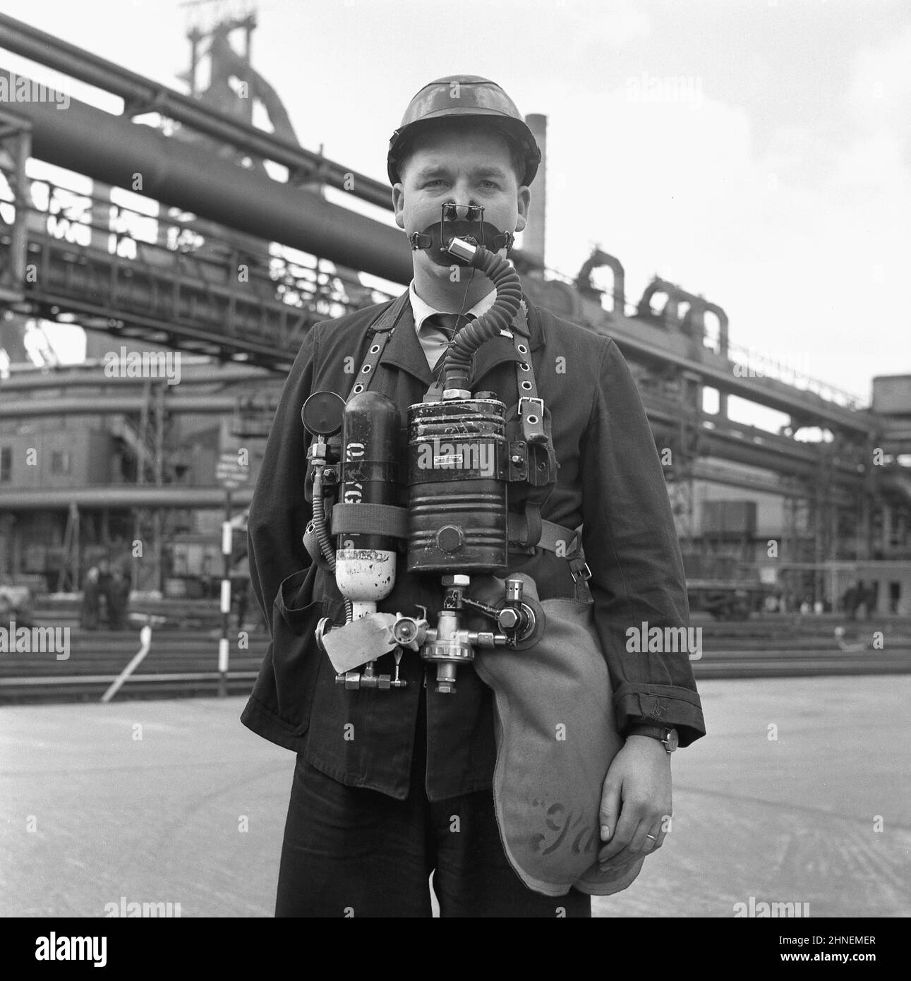 Fin 1950s, historique, en dehors d'une usine de fabrication de l'acier, un travailleur portant le dernier appareil respiratoire d'urgence - connu sous le nom de resreniflard - qui serait utilisé sur le site en cas d'explosion, rendant l'air dangereux pour la vie. Ce reniflard avait deux bouteilles d'oxygène, un ballon respiratoire et un embout buccal. De grands sites industriels, comme celui-ci à Abbey Works, Port Talbot, pays de Galles, Royaume-Uni, les plus grands aciéries d'Europe, ont dû être préparés. Banque D'Images