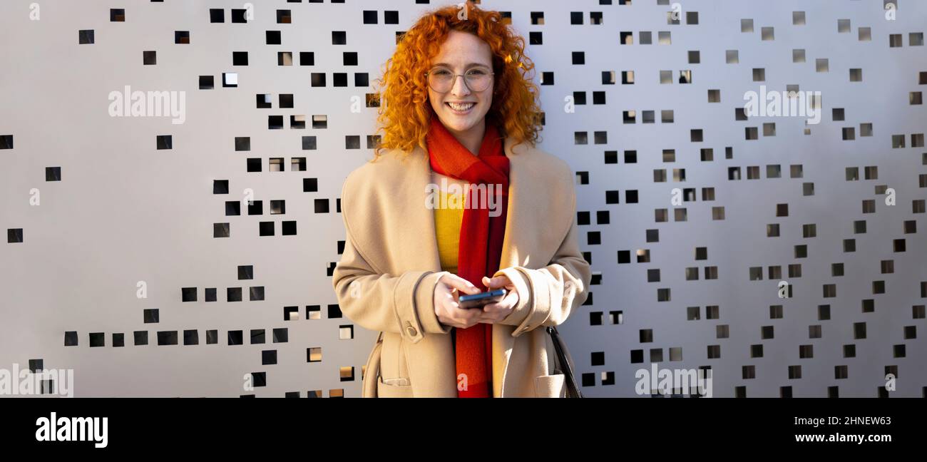 Ginger femme avec un smartphone debout devant un arrière-plan moderne Banque D'Images
