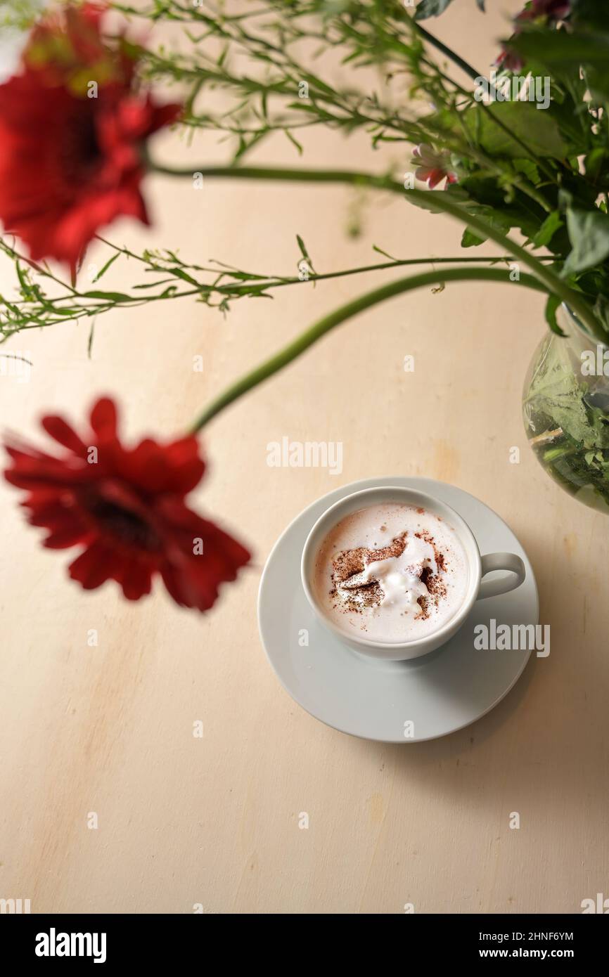Tasse avec boisson chaude au chocolat et crème fouettée sous un bouquet flou de fleurs rouges sur une table en bois clair, espace copie, vue de dessus, sélection de FO Banque D'Images