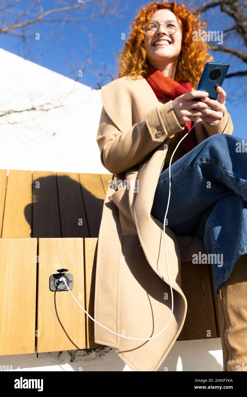 Une jeune femme à tête rouge charge son smartphone sur un banc intelligent lors d'une chaude journée de printemps Banque D'Images