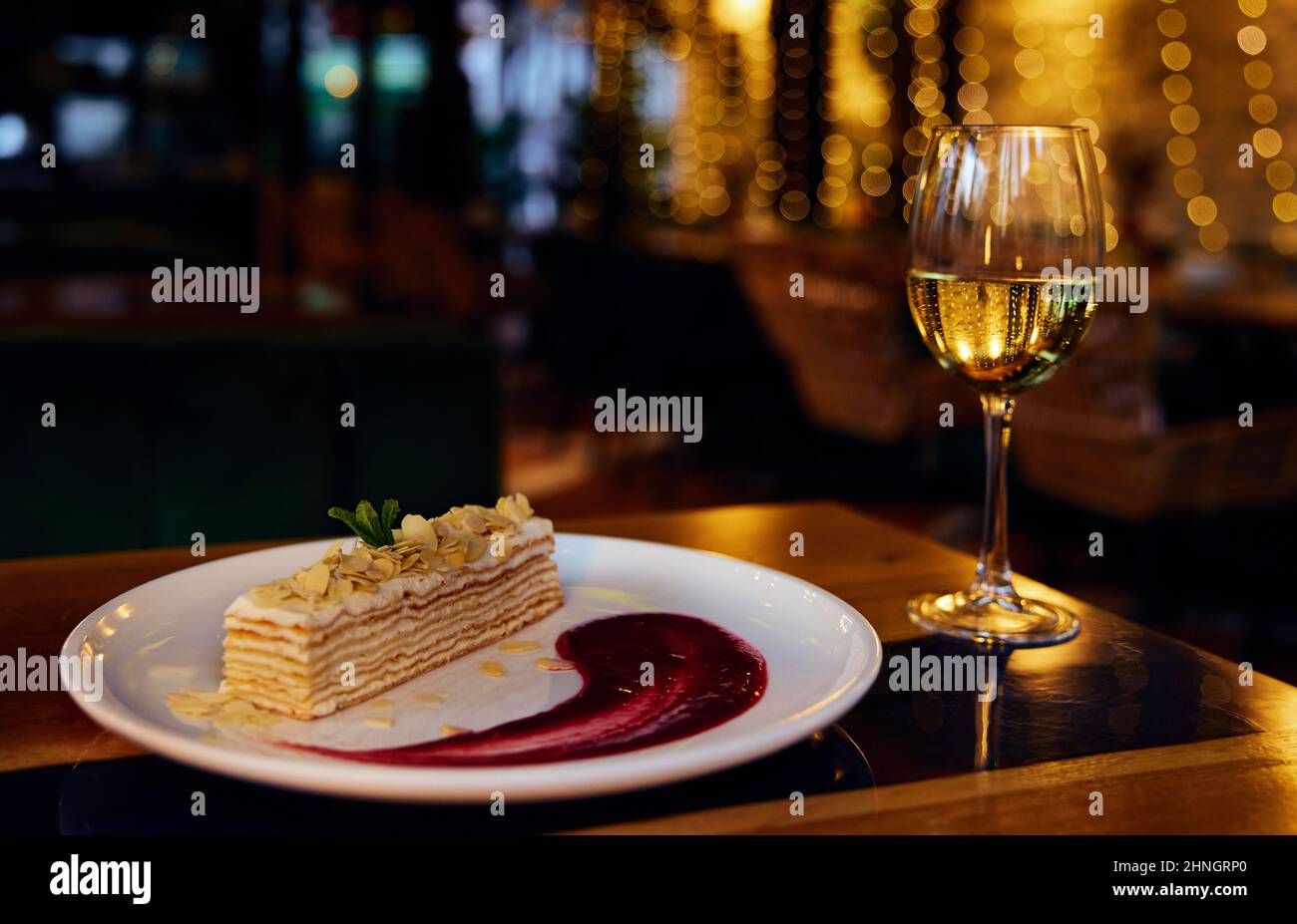 Un verre de vin blanc et un gâteau italien Napoléon et un sirop de fraise dans la plaque en céramique blanche sur une table de restaurant avec éclairage réflecte Banque D'Images