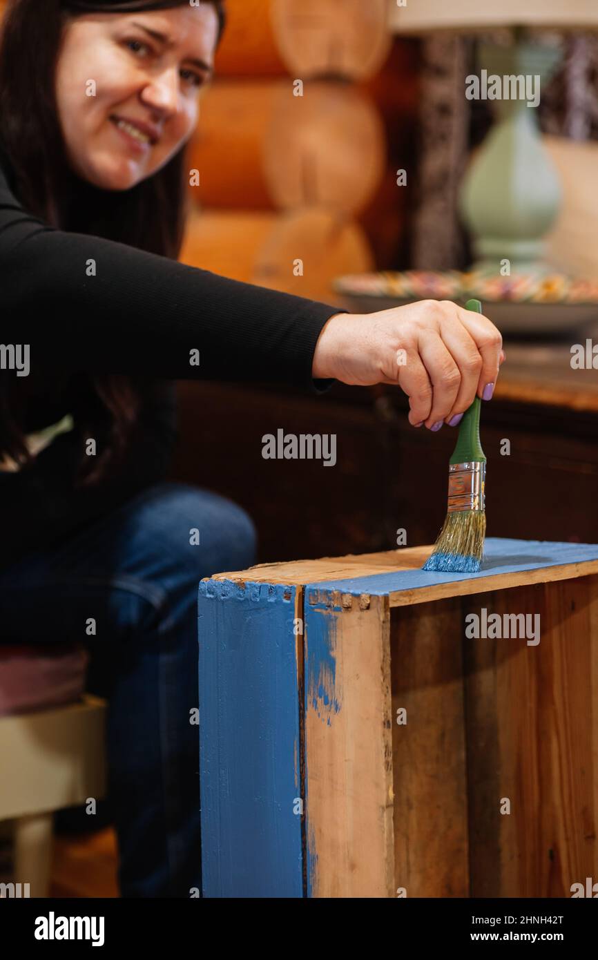 Femme d'âge moyen souriante regardant l'appareil photo avec une brosse à la main peinture super vieux tiroir de placard en bois bleu. Réutilisation des objets anciens. Durable Banque D'Images