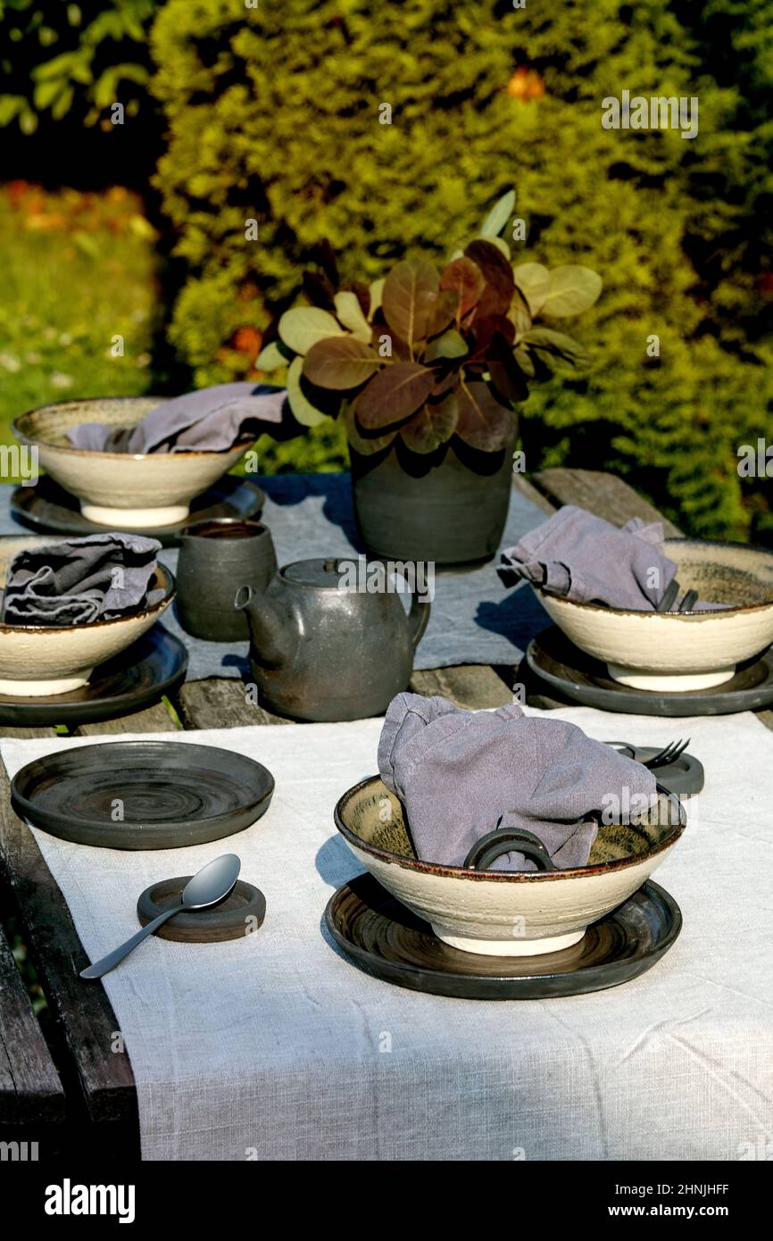 Table rustique située à l'extérieur dans le jardin avec vaisselle en  céramique artisanale vide, assiettes noires et bols rugueux, théière sur la  table en lin sur une ancienne ta en bois Photo