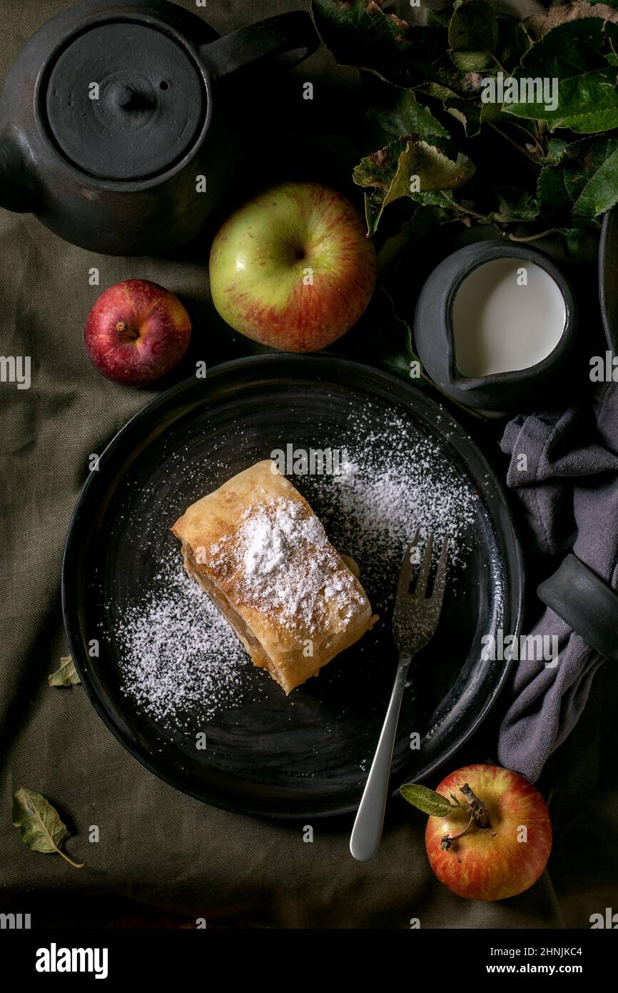 Tranche de tarte traditionnelle maison à la strudel aux pommes dans une assiette en céramique noire servie avec des pommes mûres fraîches, des branches, une fourchette et une théière sur un nappe en lin sombre Banque D'Images