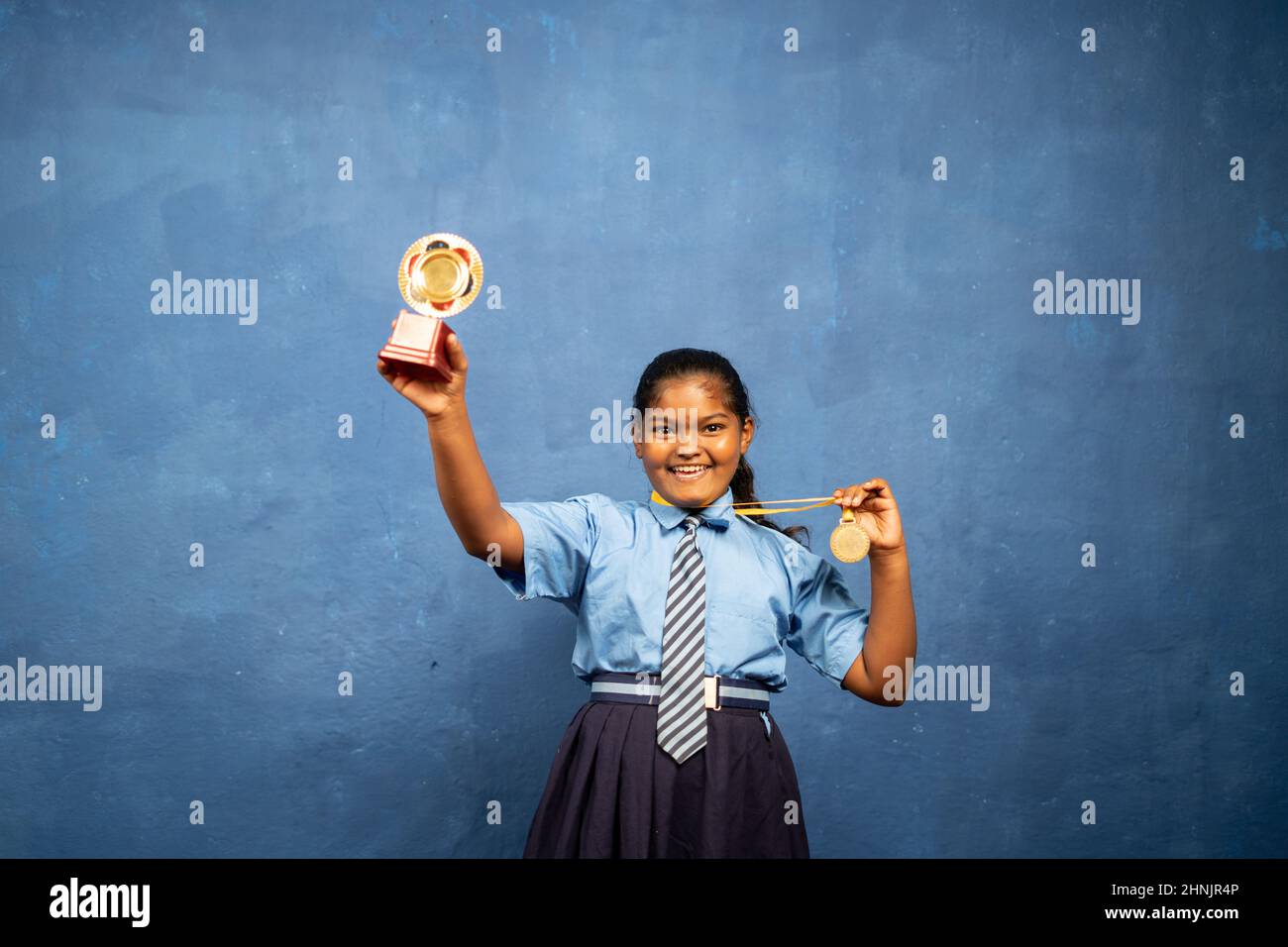 Profiter de l'école indienne jeune fille célébrer en montrant le trophée et la médaille en regardant la caméra - concept de réalisation, fier, réussi. Banque D'Images