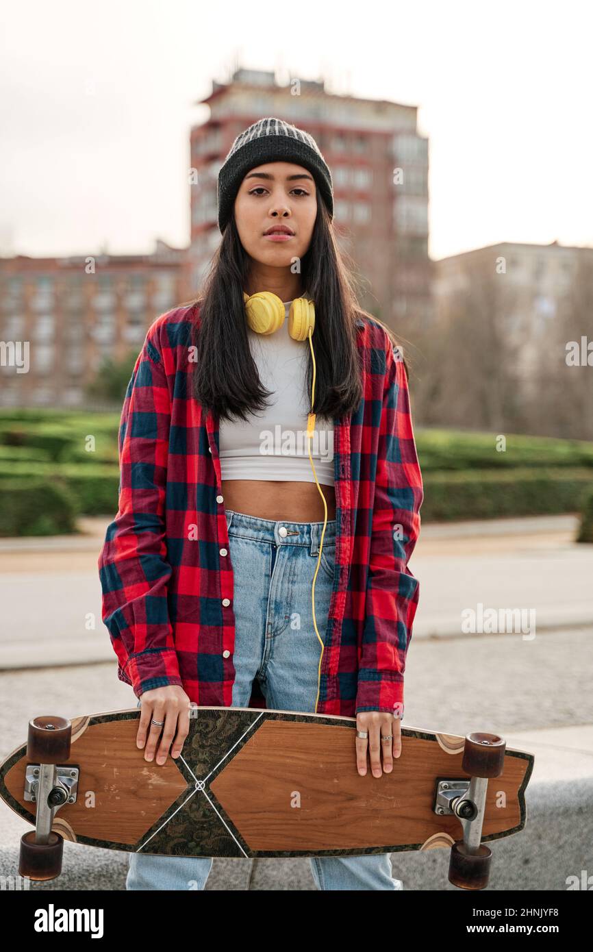 Belle jeune femme hipster posant avec un skateboard. Femme hispanique latine portant des vêtements décontractés. Banque D'Images