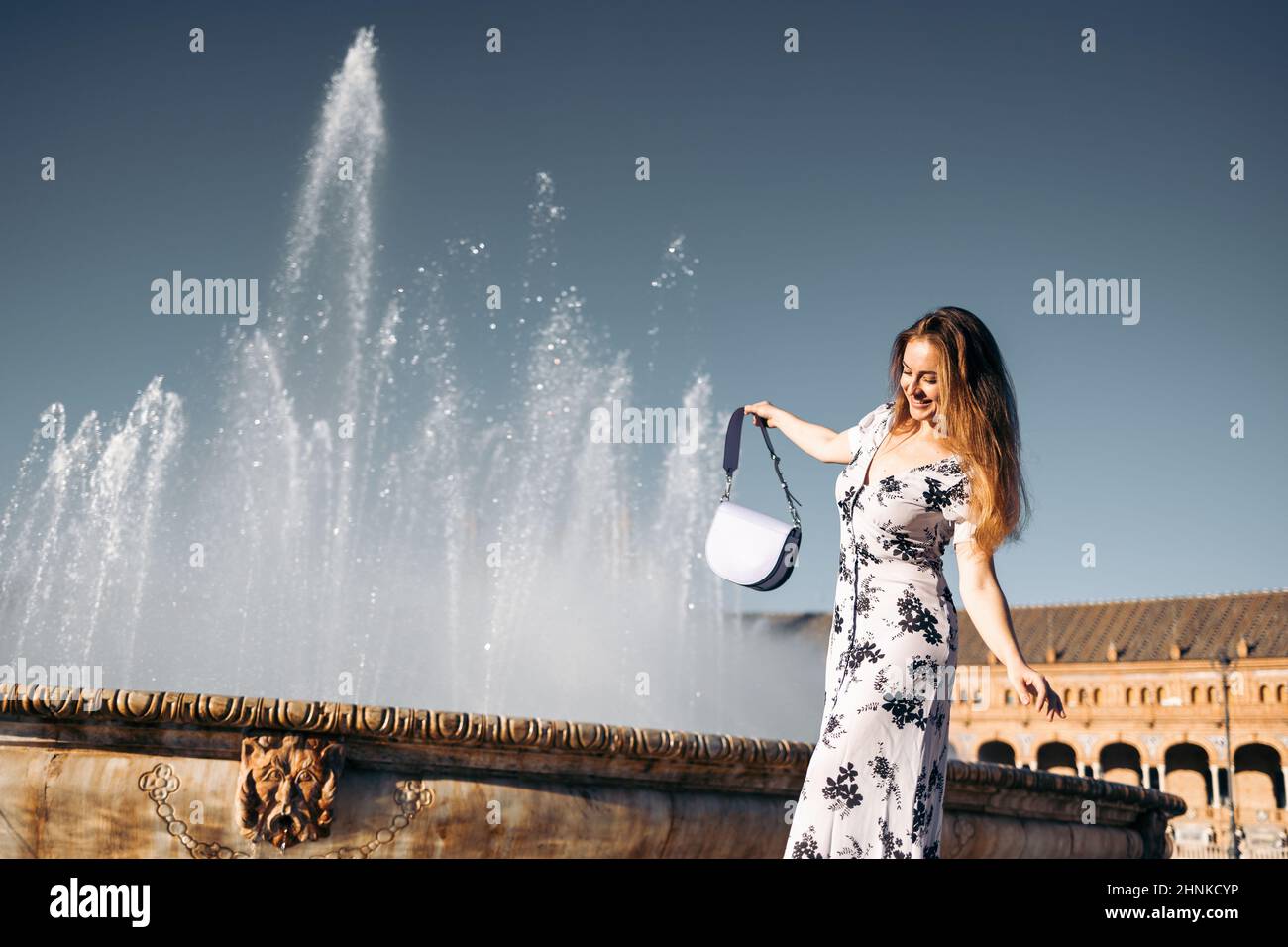 Jolie femme souriante avec sac à main et robe d'été s'amuser près d'une fontaine en Europe Banque D'Images