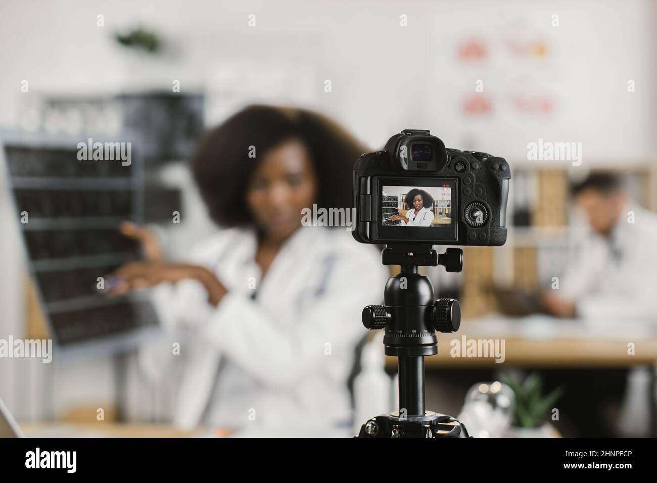 Femme africaine confiante en blouse de laboratoire blanche parlant des résultats de l'acquisition de rayons X pendant l'enregistrement de la diffusion. Mise au point sur l'écran d'une caméra vidéo moderne. Banque D'Images