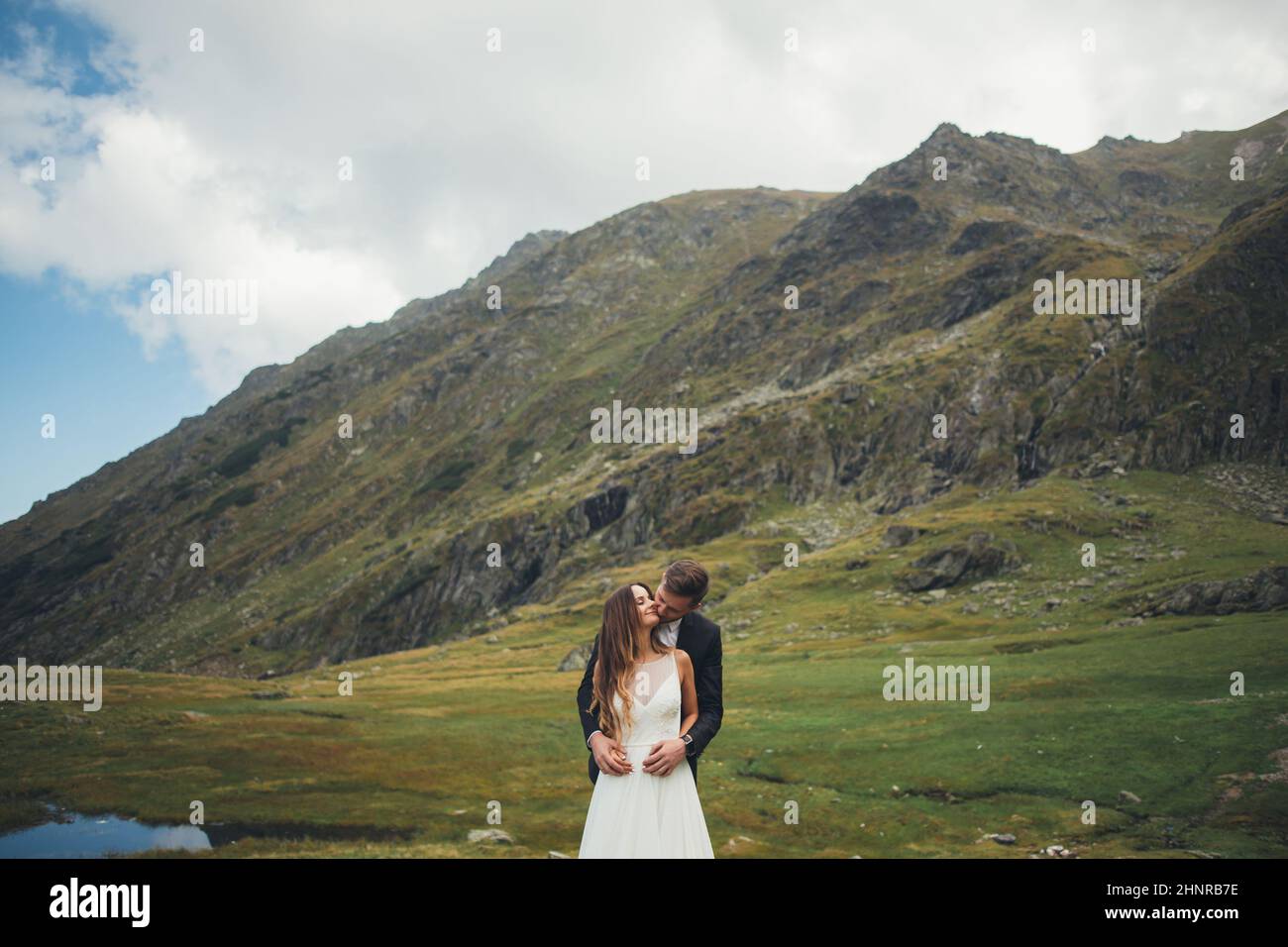 Portrait du marié embrassant et embrassant la mariée de derrière par les épaules posant sur le fond de montagnes. Portrait élégant. Famille heureuse ensemble. Banque D'Images