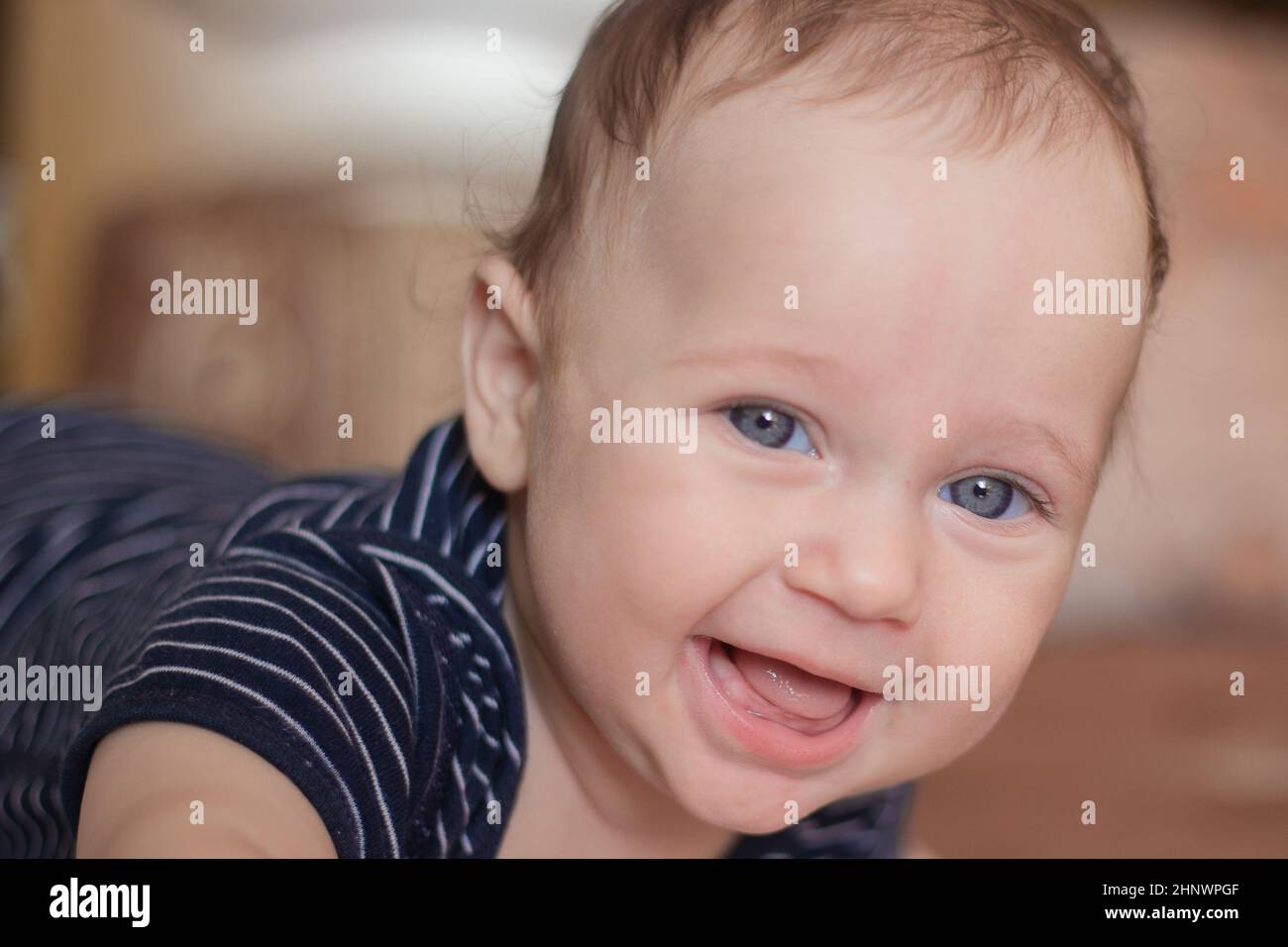 Un enfant sans dents avec de beaux yeux bleus et un front élevé d'apparence caucasienne se trouve sur son ventre et rit dans la caméra dans le dos flou Banque D'Images