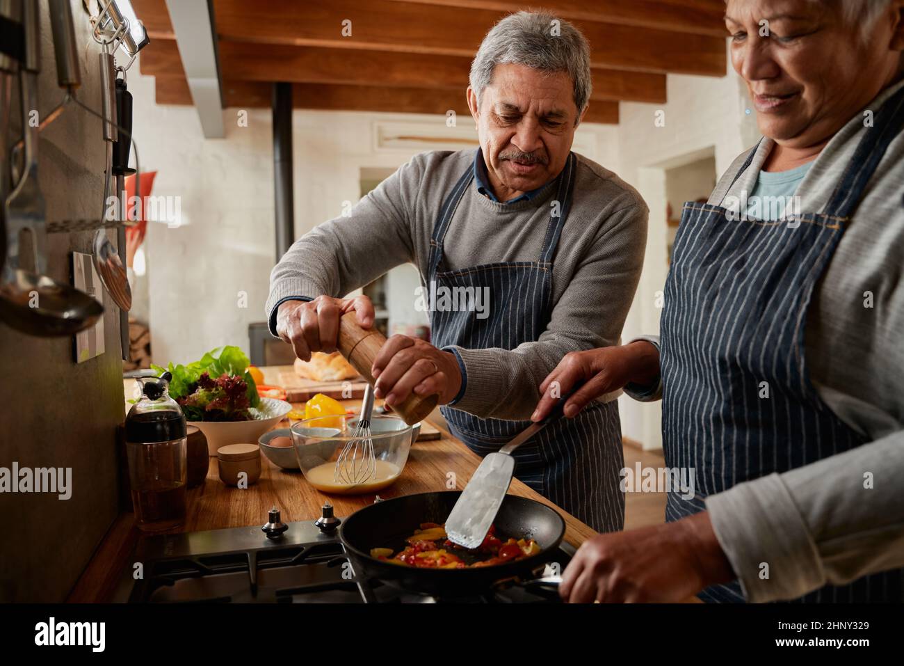 Un couple multiethnique âgé prépare le petit-déjeuner dans une cuisine moderne. Mari moulant les épices pendant que la femme remue les légumes colorés. Banque D'Images