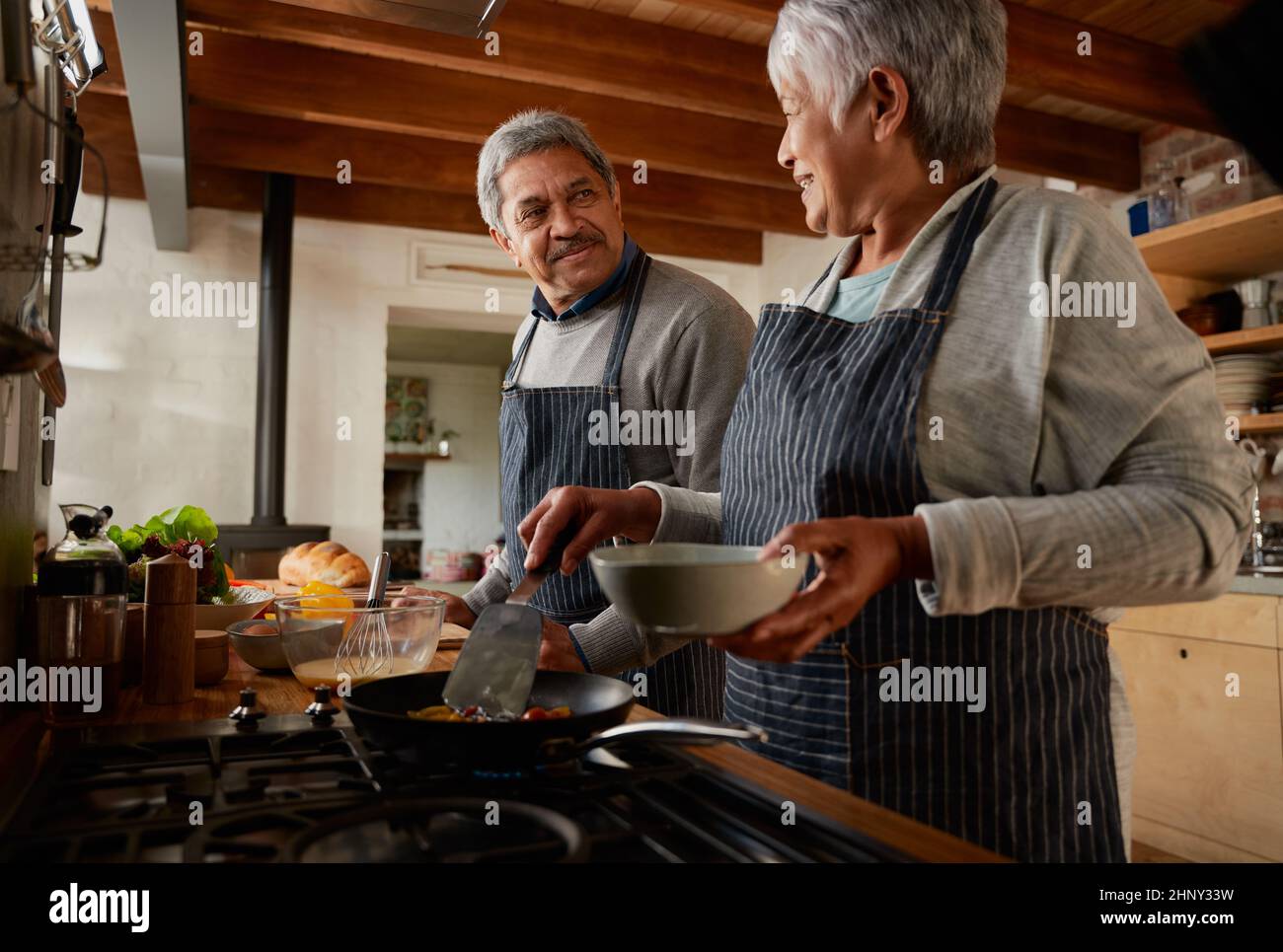 Un couple multiethnique âgé se souriant les uns les autres tout en cuisinant un petit déjeuner sain dans une cuisine moderne. Banque D'Images