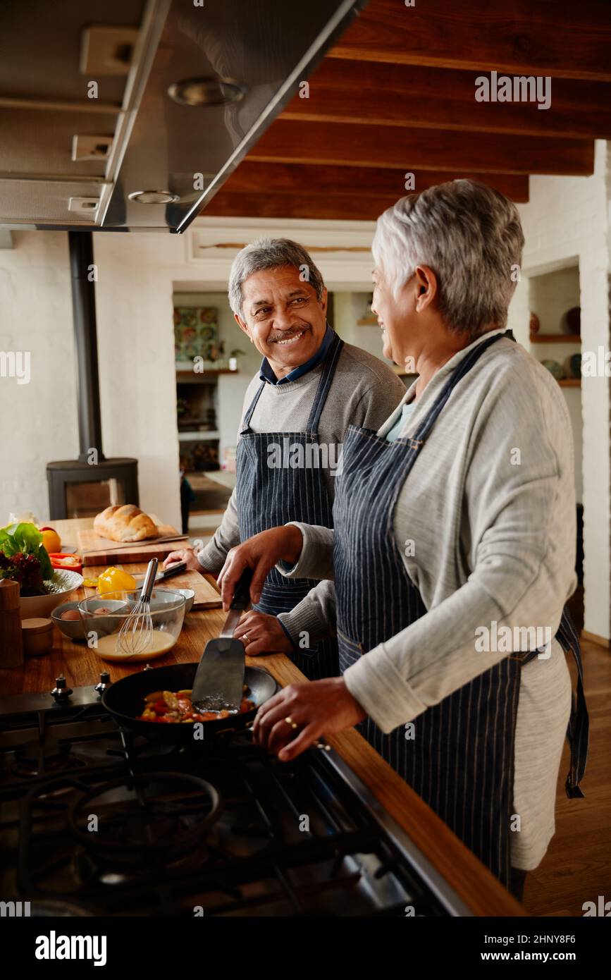 Couple multiethnique âgé regardant les uns les autres heureux tout en cuisinant le petit déjeuner dans leur cuisine moderne. Banque D'Images
