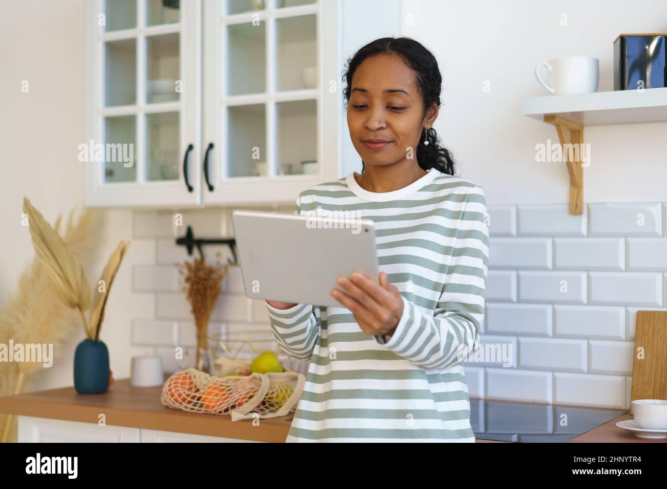 Femme afro-américaine utilisant un appareil numérique dans la cuisine. Recherche d'une recette avant de commencer la cuisson Banque D'Images