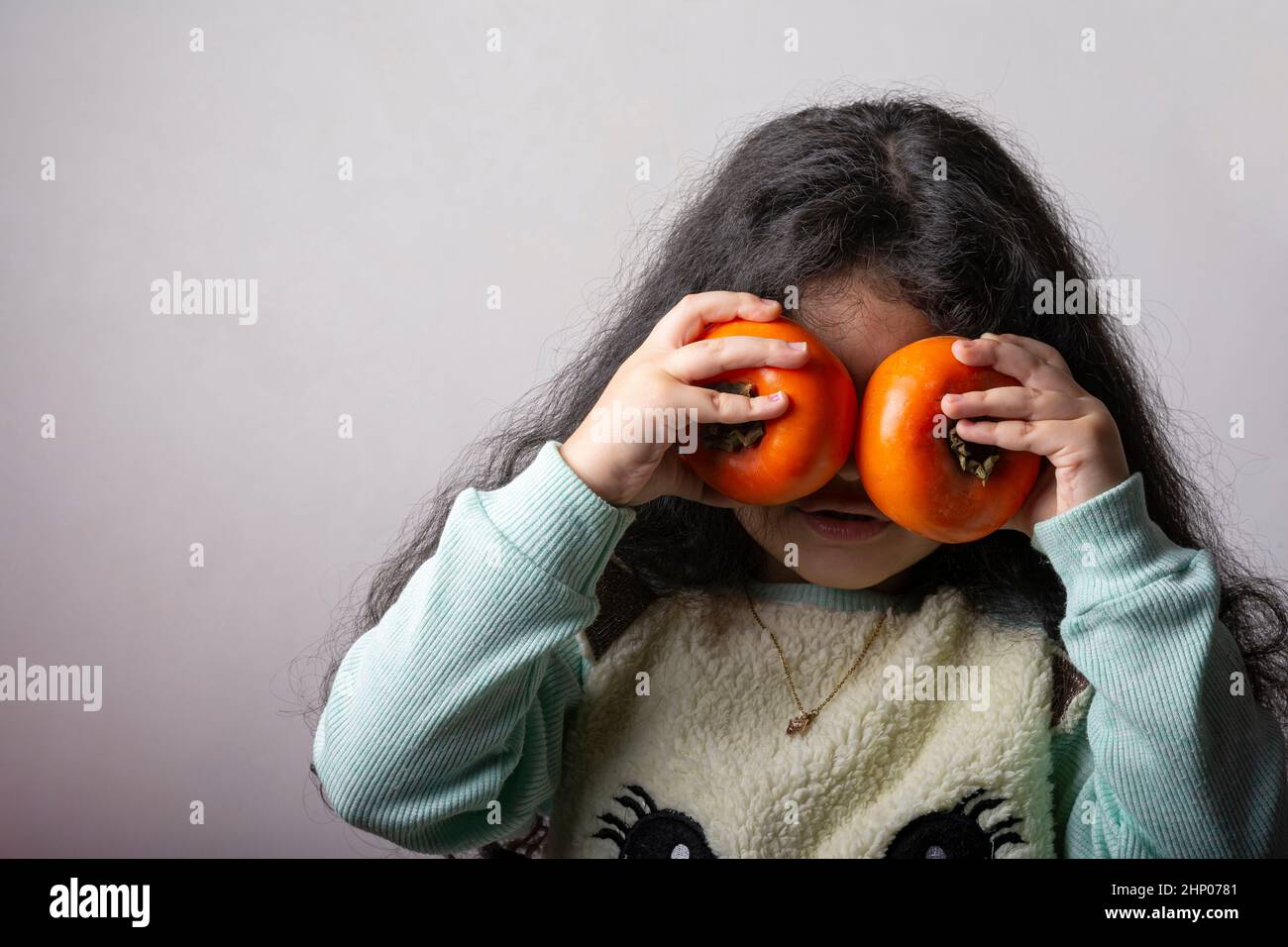 Petite fille tenant deux persimmons dans la fornt des yeux, couvre son visage. Banque D'Images