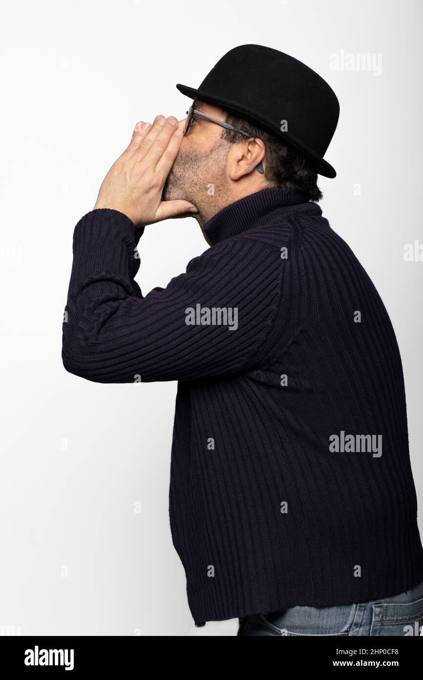 Portrait d'un homme d'âge moyen avec des lunettes, un chapeau de style bowling et des lunettes rondes. En profil, il murmure avec ses mains devant sa bouche. Isolé sur ba blanc Banque D'Images