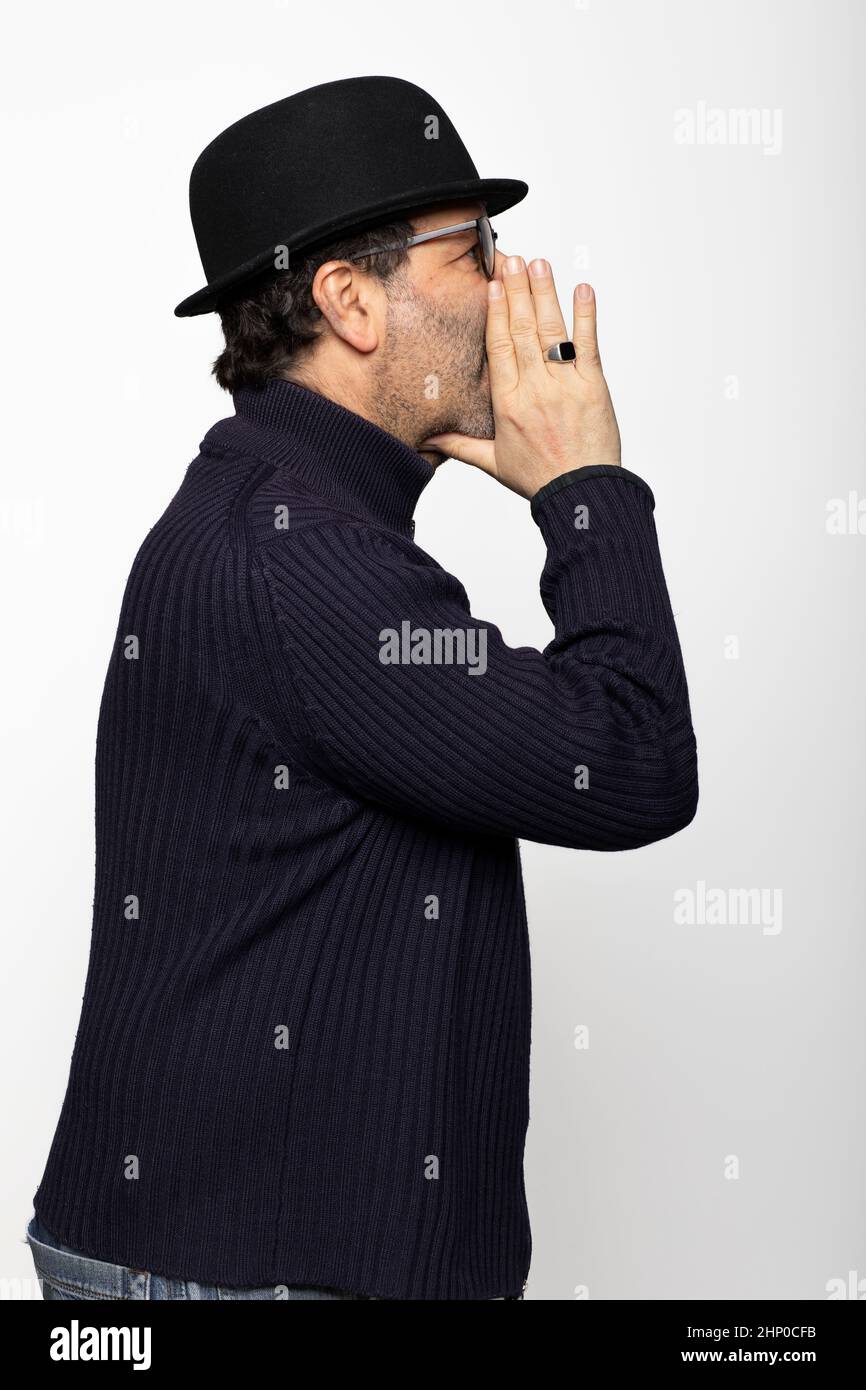 Portrait d'un homme d'âge moyen avec des lunettes, un chapeau de style bowling et des lunettes rondes. En profil, il murmure avec ses mains devant sa bouche. Isolé sur ba blanc Banque D'Images