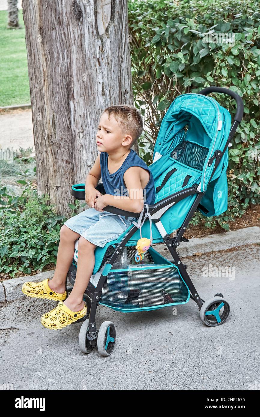 Des frondes de sourcils d'avant-garde assis dans une poussette bleue de bébé frère marchant dans le parc. Le garçon aux cheveux justes est assis et se repose fatigué de la longue promenade sur les vacances d'été Banque D'Images