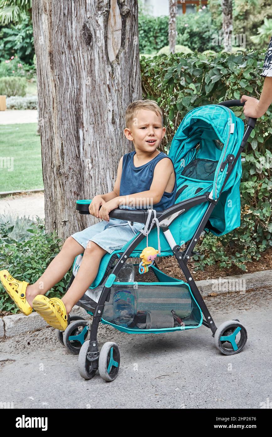 Des frondes de sourcils d'avant-garde assis dans une poussette bleue de bébé frère marchant dans le parc. Le garçon aux cheveux justes est assis et se repose fatigué de la longue promenade sur les vacances d'été Banque D'Images