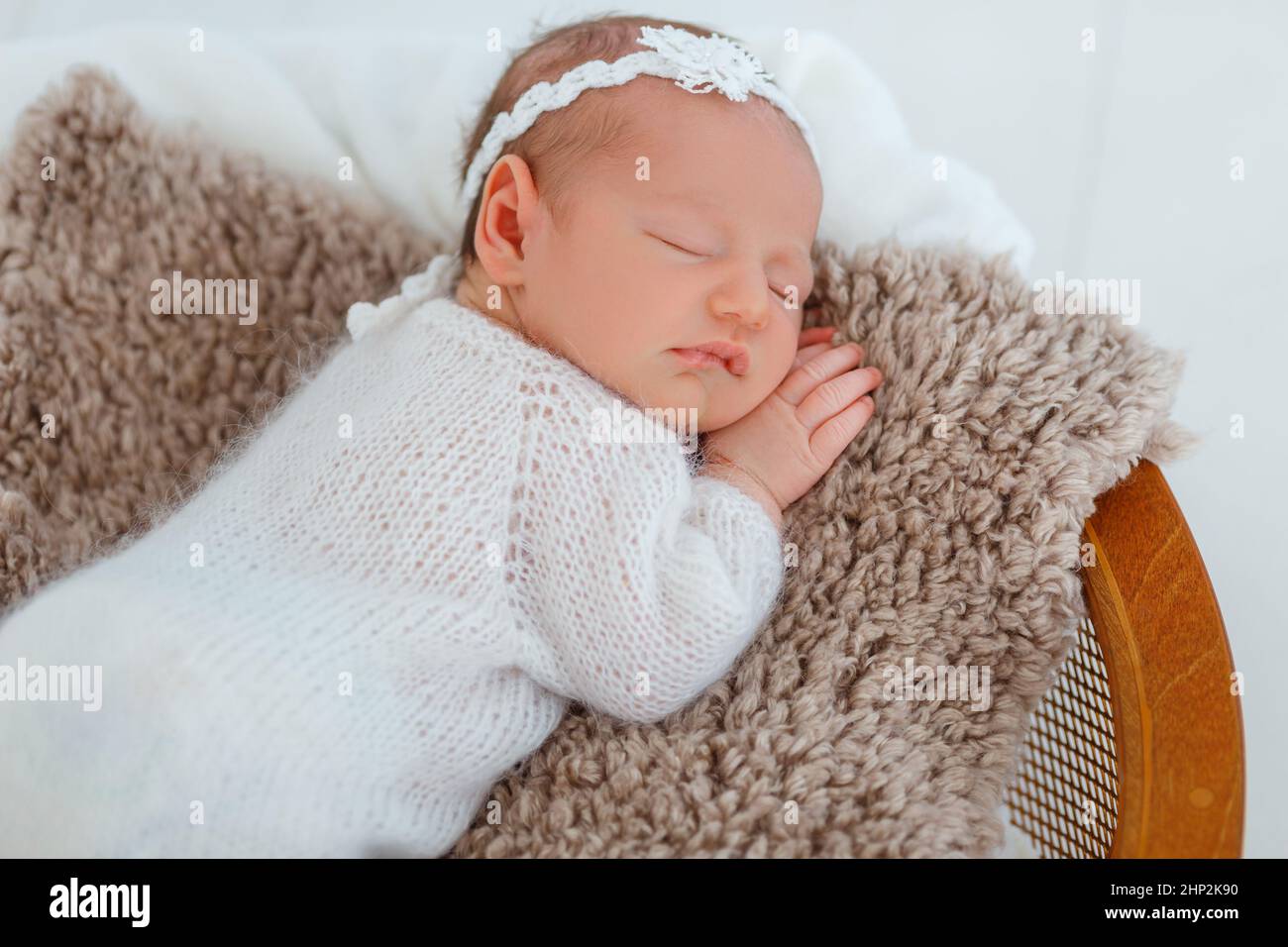 Nouveau-né en costume blanc tricoté dormir dans un panier en bois. Enfant se reposant dans une posture mignonne. Petite fille avec serre-tête tendre sur la tête. Photogr. Nourrisson créatif Banque D'Images