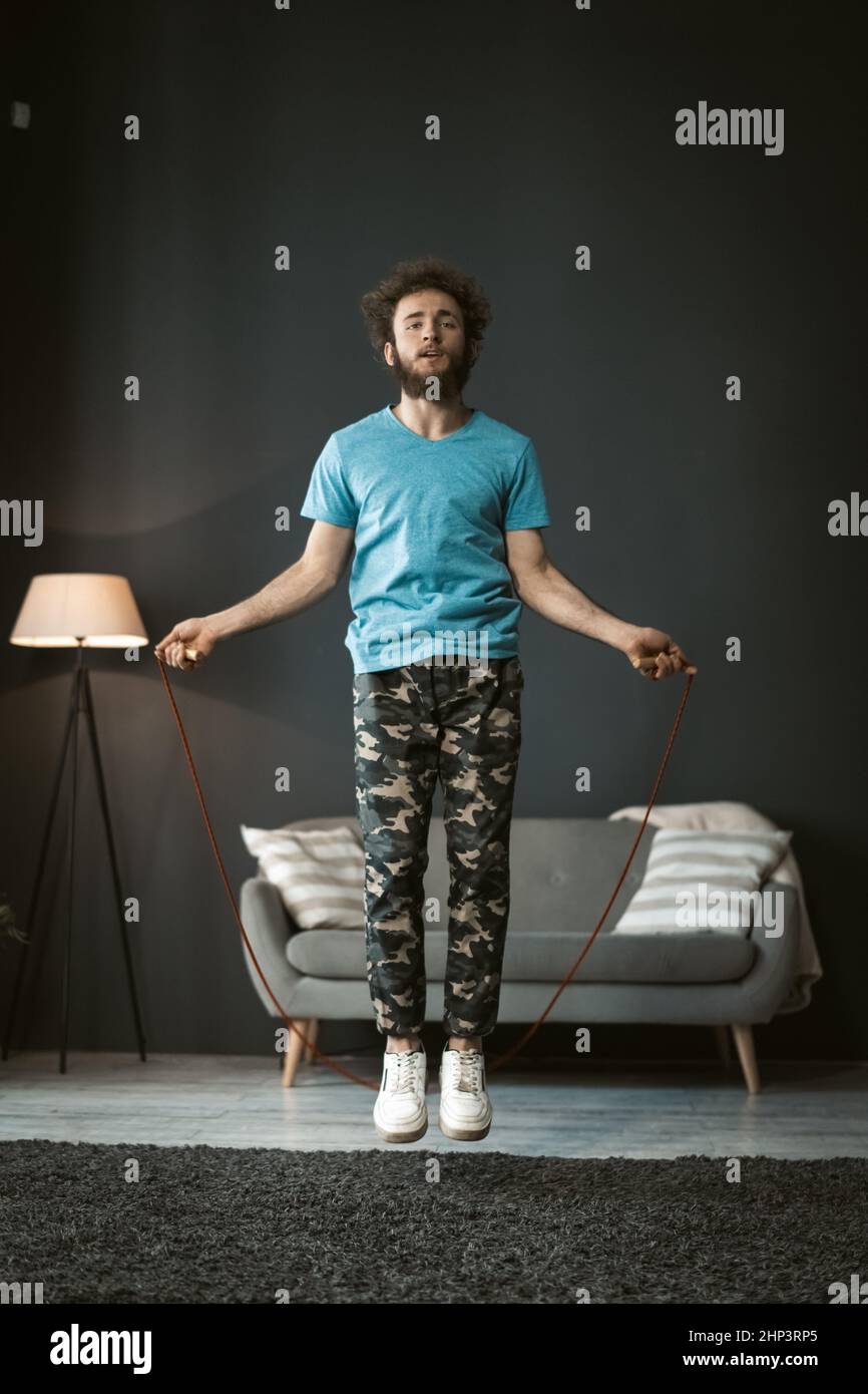 corde sautant un jeune homme qui fait de l'activité à la maison. Jeune homme en auto-isolement ou en quarantaine, jours de pandémie. Beau cheveux bouclés homme utiliser la corde à sauter pour rester en forme et en bonne santé. Banque D'Images