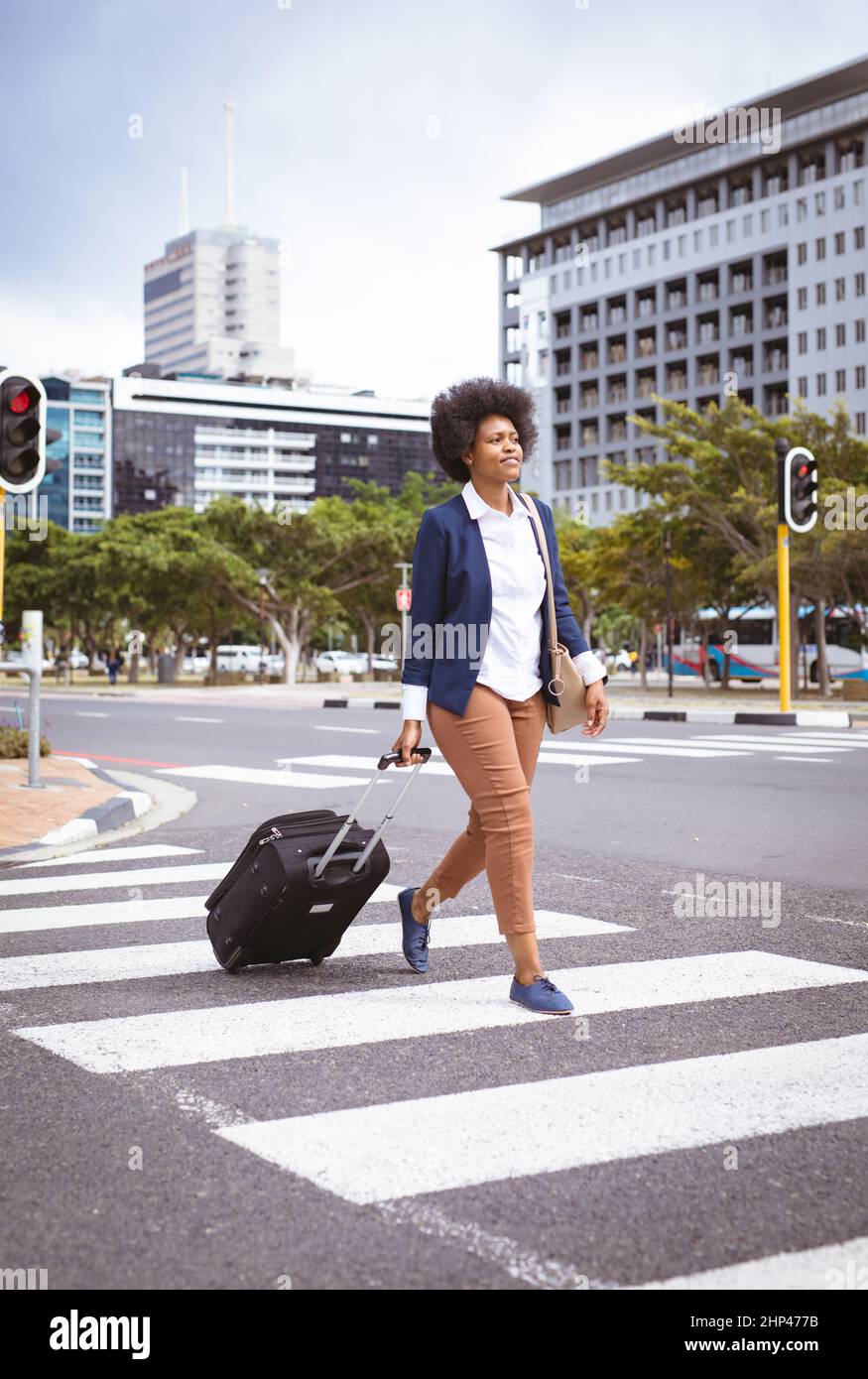 Femme d'affaires afro-américaine de taille moyenne avec bagages à roulettes et porte-monnaie traversant des passages de côté en ville Banque D'Images