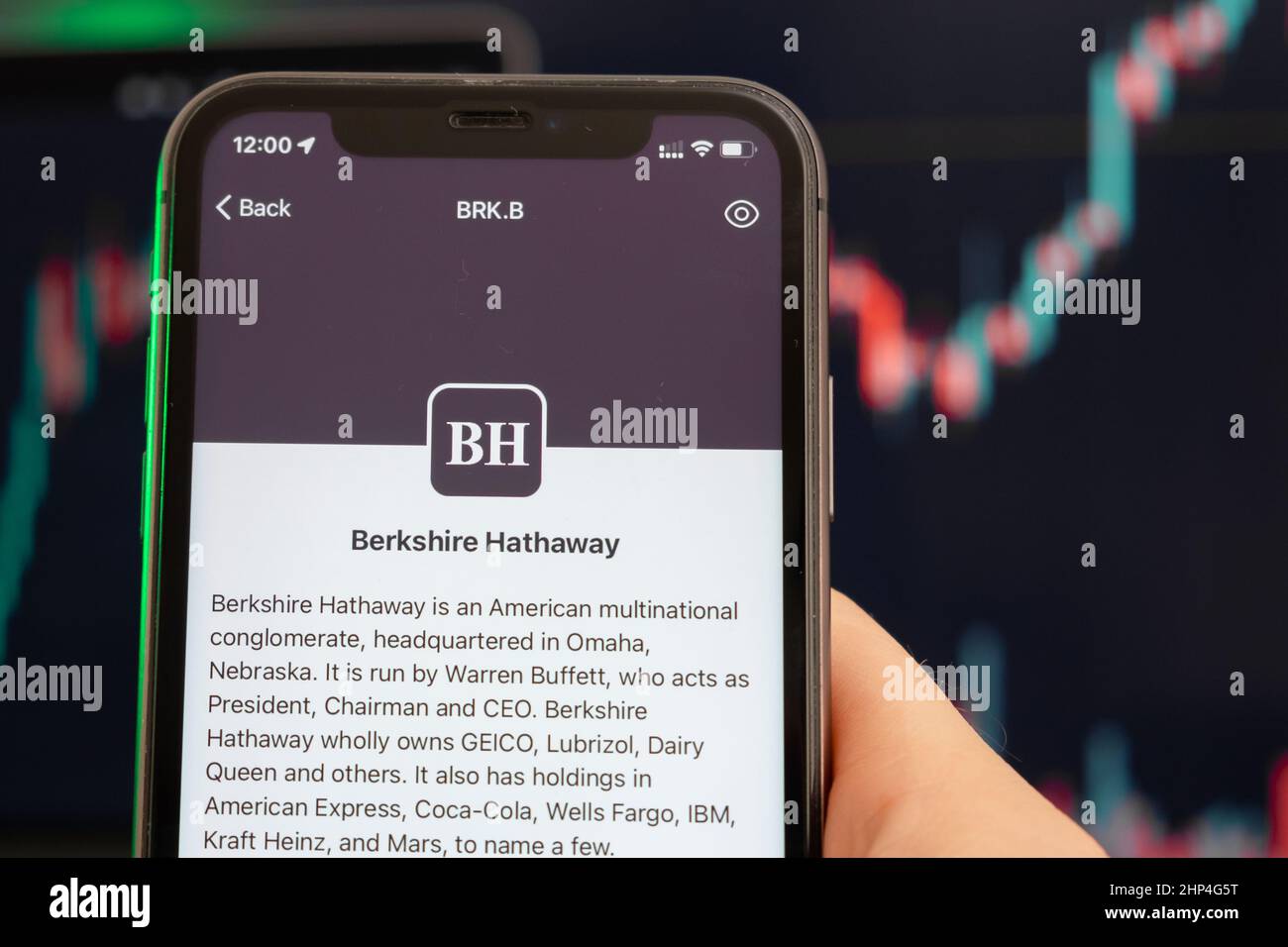 Berkshire Hathaway BH prix de l'action tendance avec des graphiques sur le fond. Homme mains tenant un smartphone avec logo sur l'écran, février 2022, San Francisco, Etats-Unis. Banque D'Images