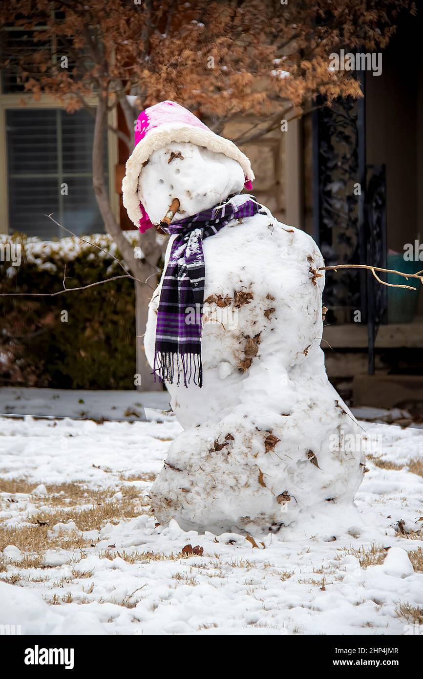 Bonhomme de neige avec chapeau rose et foulard violet et vieux cigare dans sa bouche debout devant la maison. Banque D'Images