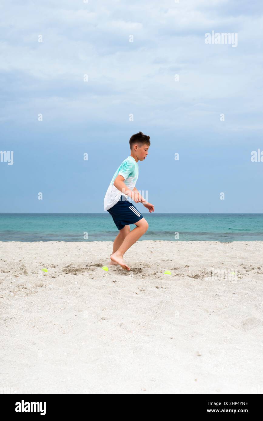 Le garçon s'entraîne sur le sable au bord de la mer. Entraînement sur les jambes. Préparation générale de l'allumage. Banque D'Images