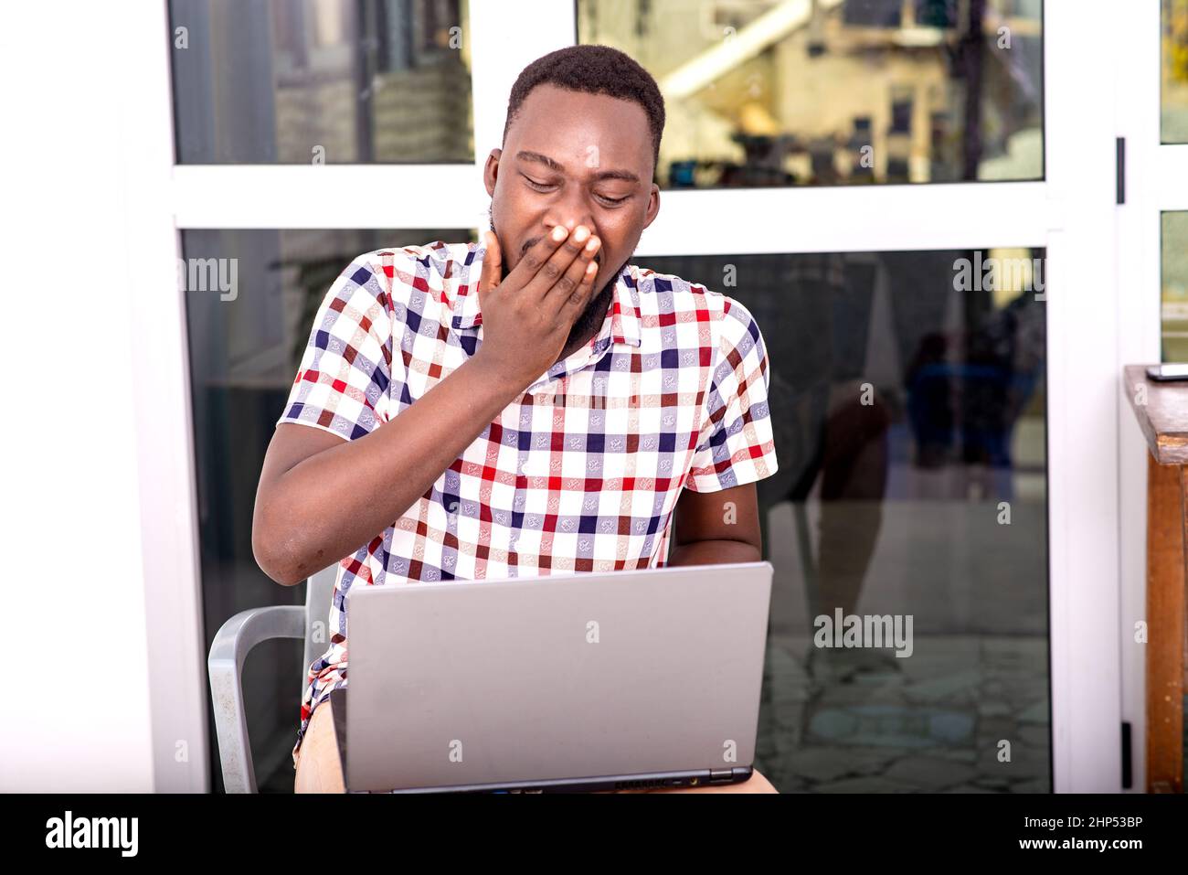 jeune homme d'affaires en chemise à carreaux assis sur une chaise au bureau avec un ordinateur portable sur les cuisses bâillant avec la main sur la bouche. Banque D'Images