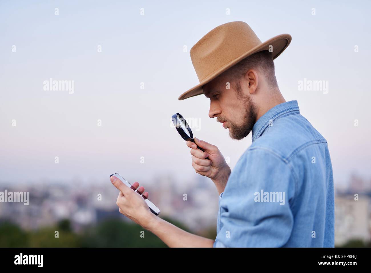 Beau barbu qui regarde avec la loupe sur le smartphone. Homme portant un Jean chemise et chapeau regardant avec la loupe au téléphone mobile comme détective ou enquêteur concept. Image de haute qualité Banque D'Images