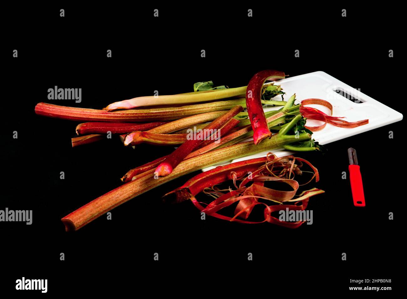 Disposition de la tige de rhubarbe sur le plan de cuisine, peau raclée et éplucheur sur fond noir. Gommage de la rhubarbe. Banque D'Images