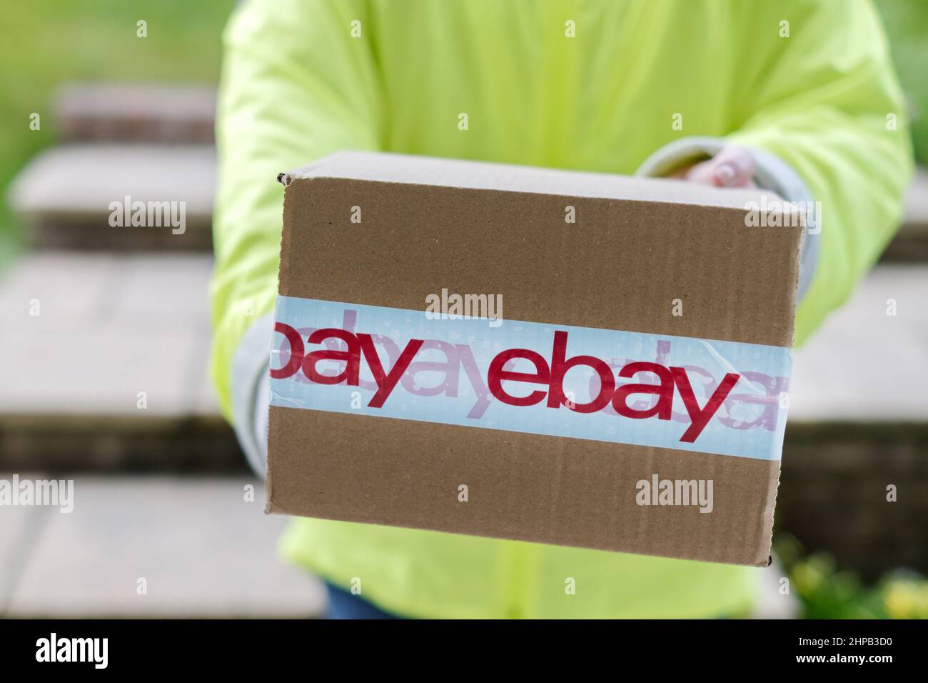 Un coursier livrant un colis eBay à la porte d'entrée d'un client. La boîte en carton est enveloppée de ruban adhésif montrant clairement le logo eBay. Banque D'Images