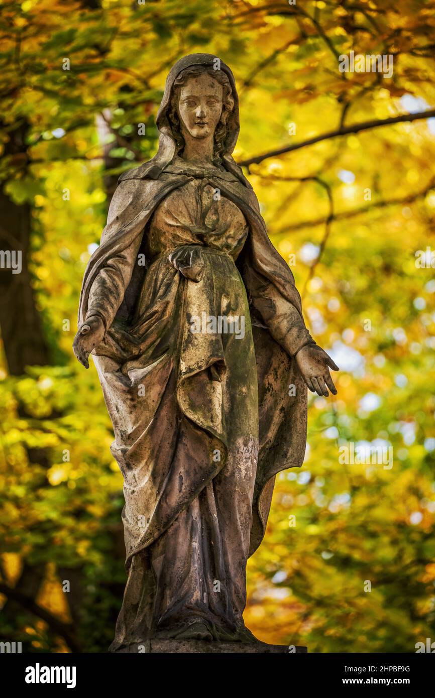 Statue de dame à capuchon avec bras ouverts dans la robe classique et l'expression aimable, ancienne tombe sculpture en pierre contre le feuillage d'automne dans le cimetière. Banque D'Images