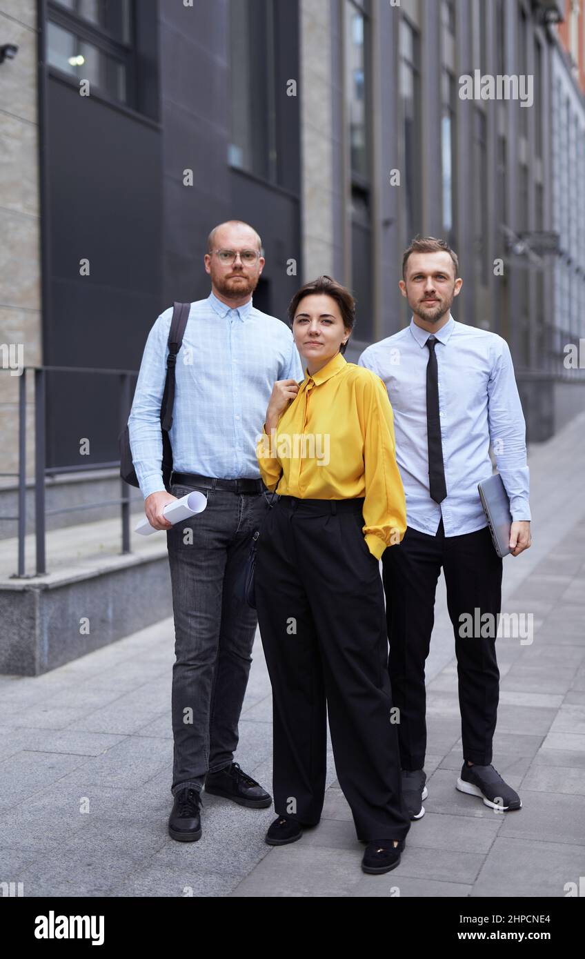 Jolie femme caucasienne confiante dans un chemisier jaune et deux hommes regardant la caméra souriant. Portrait de moyenne prise de vue d'une équipe d'affaires professionnelle réussie se posant à l'extérieur dans le centre-ville. Photo de haute qualité Banque D'Images