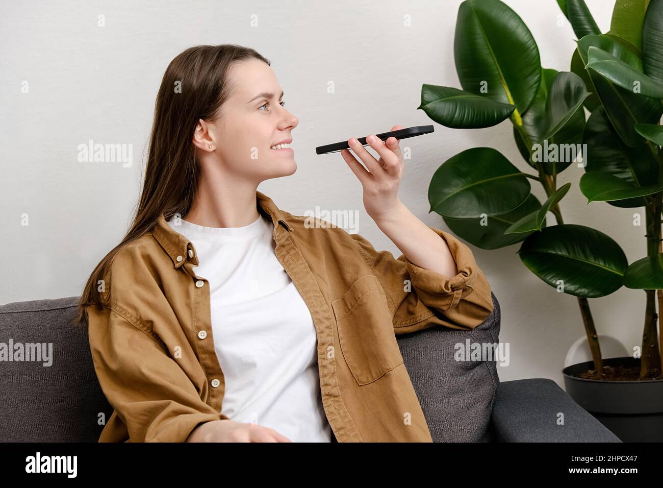 Bonne jeune femme brune assise sur un canapé à l'aide d'un smartphone tient près de la bouche parlant avec un petit ami sur le haut-parleur. Utilisation de la messagerie vocale Banque D'Images