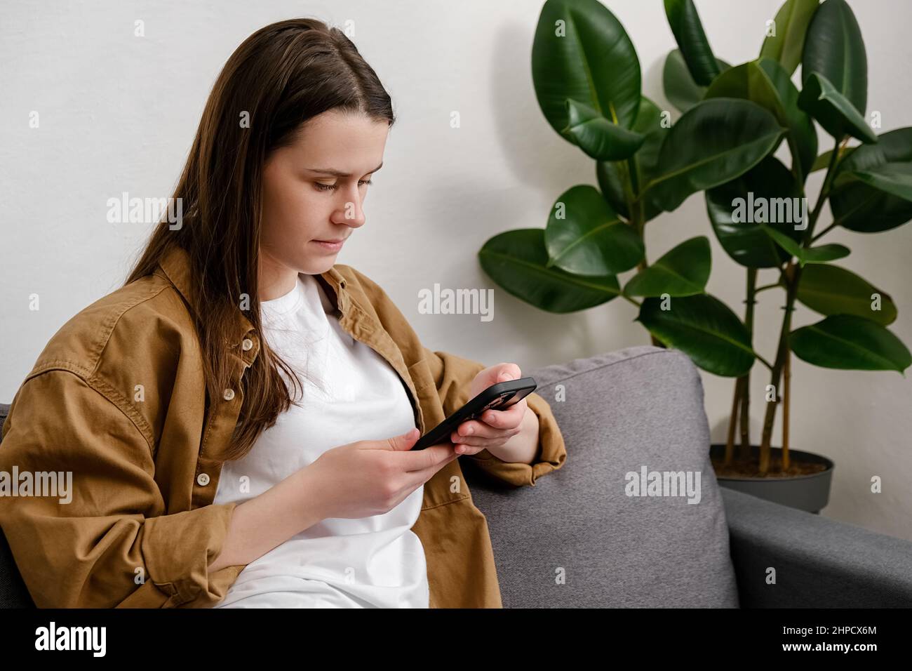 Une jeune femme calme passe du temps libre à la maison s'asseoir sur un canapé confortable dans le salon pour envoyer des SMS dans une application mobile sur un smartphone. Banque D'Images