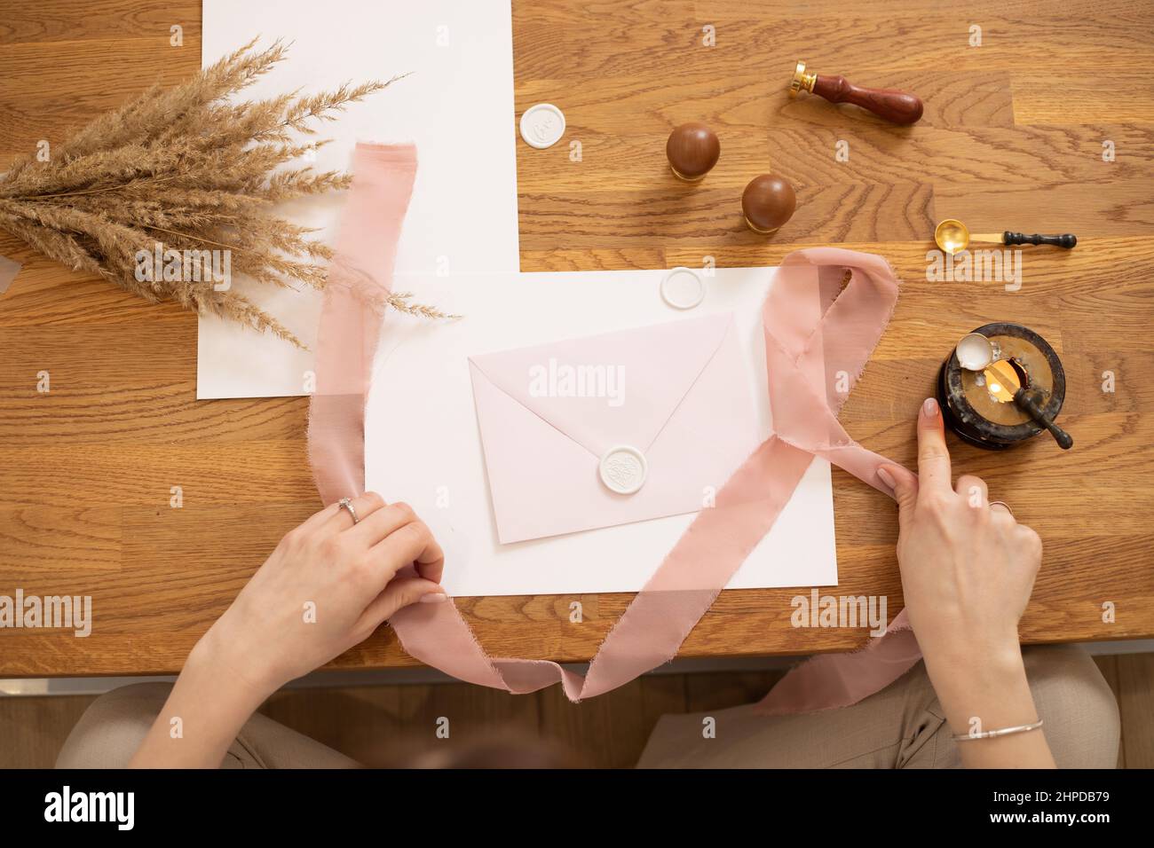 Photo rognée des mains de femme faisant l'enveloppe de message d'accueil artisanat, décorer avec ruban rose et estampage. Herbes sèches sur le bureau Banque D'Images