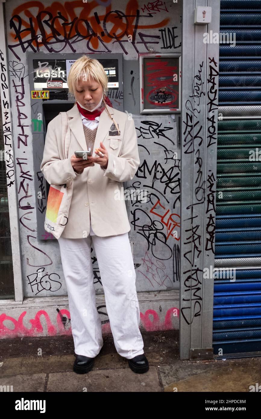 Une femme asiatique vérifie son téléphone tout en fume une cigarette. Derrière se trouve un cash point très gratitisé. Photo prise dans East Londons Brick Lane. Banque D'Images
