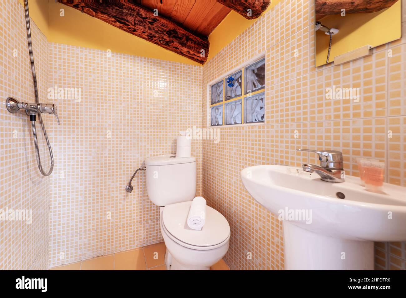 Salle de bains avec douche murale, lavabo en porcelaine blanche, miroir mural carré, carrelage en mosaïque jaune et poutres apparentes Banque D'Images