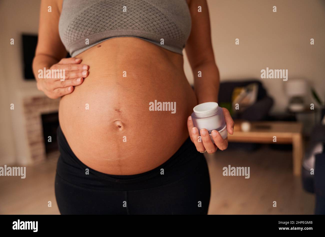 Torse de femme caucasienne enceinte debout, appliquant de la crème sur son ventre. Un style de vie actif et heureux dans une maison moderne en journée. Banque D'Images